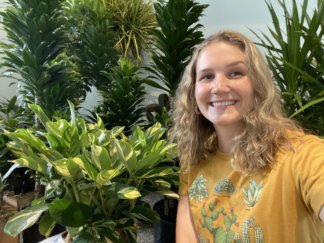 Emma Hondzinski standing next to indoor houseplants wearing a shirt with plants printed on it